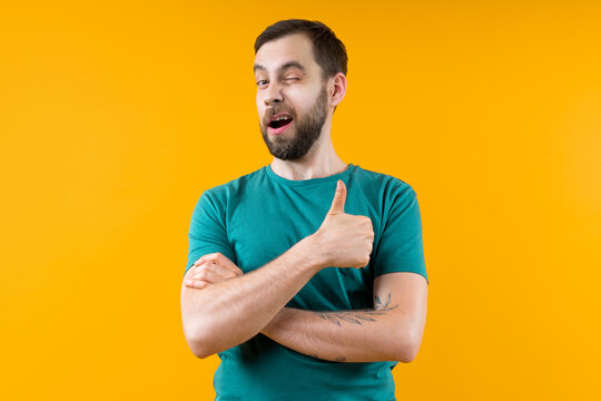 Studio portrait of confident young man posing over bright colored yellow background winking and holding thumb up