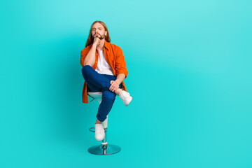 Full length photo of intelligent cool minded man with long hairdo dressed orange shirt sit on chair isolated on teal color background