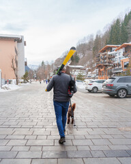 A person with skis on their shoulder walks a dog in a snowy mountain town, embodying the winter sports lifestyle. This image can be a concept for vacation, leisure, and the joy of winter.