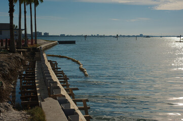 Fototapeta premium Medium view from Gulfport ,Florida Marina looking west with s curve of concrete seawall being repaired towards Boca Ciega Bay in afternoon sun. Green Palm trees on the side , calm water and blue sky.