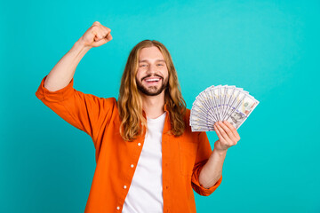 Portrait of ecstatic man with long hairstyle dressed orange shirt win big money in lottery fist up...
