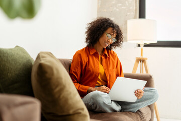 Smiling attractive African American woman using laptop computer, typing, communication, shopping online sitting on comfortable in office. Successful happy freelancer, programmer working from home 