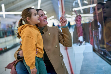 Mother and daughter gaze at a store window in the mall. The mother points at something, sharing a special find during their shopping adventure.