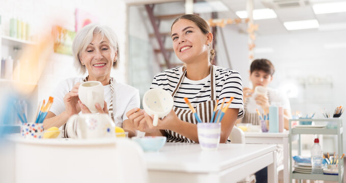 Cheerful elderly woman and young girl, participating in pottery masterclass, sitting together at table, engaging in friendly conversation while painting ceramic dishware