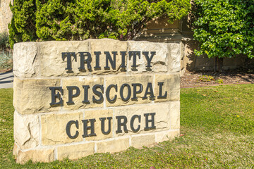 Fototapeta premium Santa Barbara, CA, USA - November 30, 2023: Trinity Episcopal Church sign set in black metal letters on beige rocky stones. Green Front lawn with hedge