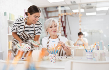 Friendly smiling young female ceramicist teaching elderly woman art of ceramic glazing and painting in modern pottery studio..