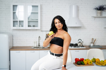 Young beautiful woman is sitting on the table in the kitchen and eating apple.