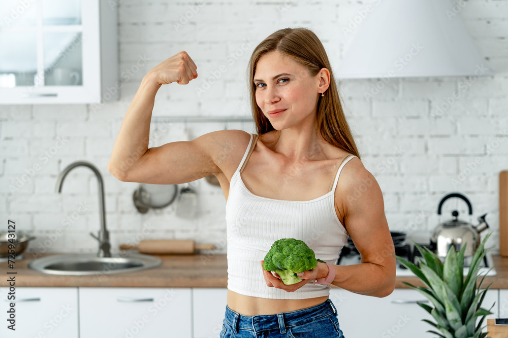 Canvas Prints Strong woman is holding broccoli and flexing her biceps in the kitchen.