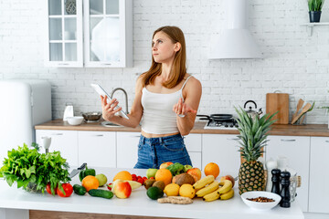 Young woman standing in the kitchen with smartphone and fresh fruits