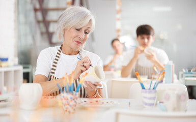 Obraz na płótnie Canvas Talented senior female artisan in apron working on embellishing ceramic mug at table in modern pottery studio, applying design with paints..