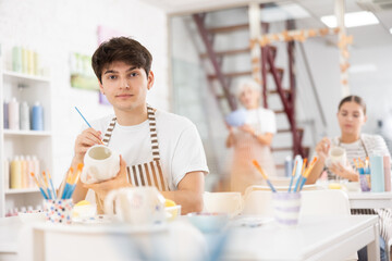Focused young guy attending pottery class, enthusiastically painting on ceramic cup, creating...