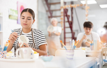 Positive young girl attending pottery class, enthusiastically painting on ceramic mug, creating...