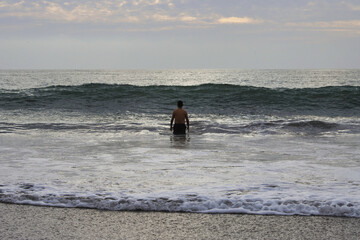 Hombre solitario mira tranquilo como viene una ola hacia el, en un día nublado al atardecer.