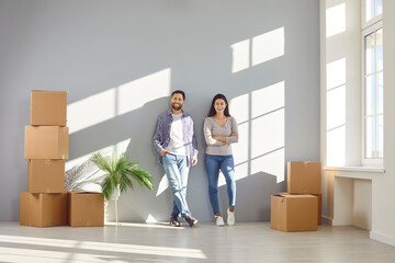 Fototapeta na wymiar Happy family couple in casual clothes are standing together near wall in rays of sunlight among cardboard boxes. Married smiling couple buys or rents new house. Real estate, relocation, own housing.