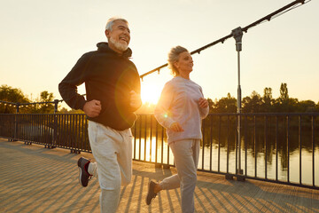 Sporty senior smiling couple in sportswear jogging along a bridge in park having sport workout at...