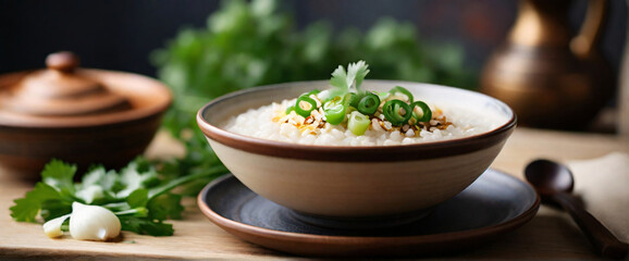 A steaming bowl of rice porridge, placed centrally on a textured, earthenware dish. The porridge is garnished with a sprinkle of chopped spring onions, a few slices of ginger, and a light drizzle