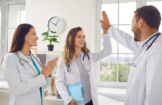 Happy smiling doctors rejoicing at success in treatment giving high five each other standing in clinic. Successful team of young medical staff in white uniform in hospital. Health care concept.