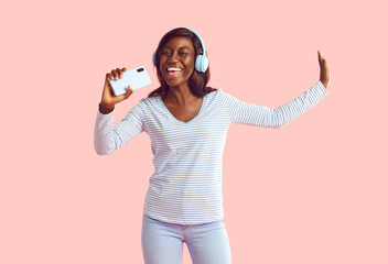 Happy cheerful joyful positive young African woman on pastel studio background uses mobile phone and noise cancellation headphones head set, listens to music, has good time, enjoys her favorite tracks - obrazy, fototapety, plakaty