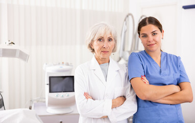 Confident senior woman cosmetologist and her assistant nurse looking at camera in clinic of aesthetic medicine