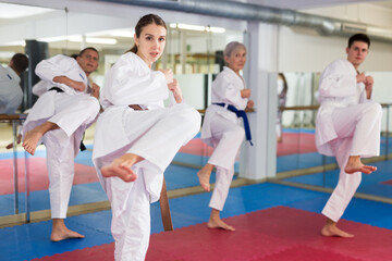Fototapeta na wymiar Various aged men and women in white kimono and belts training kata moves in gym.