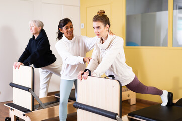 Personal female trainer controlling movements of young woman doing pilates on reformer in fitness...