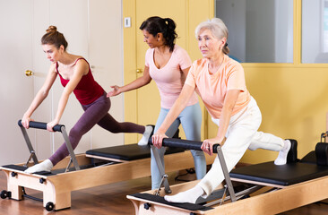 Fototapeta na wymiar Two active women practicing Pilates in a fitness studio perform an exercise using a reformer bed, where a female instructor ..helps them do it correctly