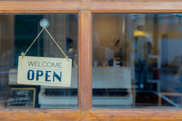 Brown wooden sign board with word "Welcome we are OPEN please come in" hanging in front of entrance door, Glass window of Restaurants, Cafe' or Shops, Business and financial background.