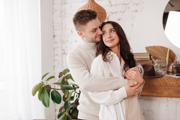 Happy young couple in love is hugging gentle and smiling together celebrating Valentine's day at cozy Scandinavian style home. Man and woman enjoying spending time together, relaxing on date.