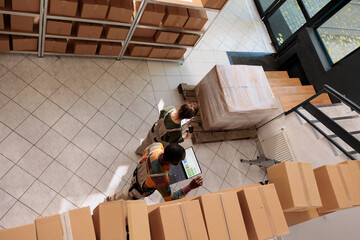 Diverse employees tracking clients online orders, checking warehouse logistics using laptop computer. Storage room supervisor preparing packages for delivery with manager. Top view concept