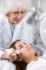 Careful old woman doctor conducting vacuum cleaning of young woman patient's face in aesthetic clinic