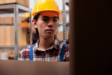 Warehouse employee in yellow helmet searching parcel on shelf while working in storage room. Young...