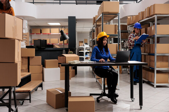 Shipment manager in yellow helmet supervising warehouse operations on laptop. Young asian woman postal service employee using parcel tracking system on computer while working in storehouse