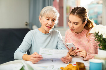Mother and daughter sitting at table and calculating finances. Daughter using her smartphone for online payment.