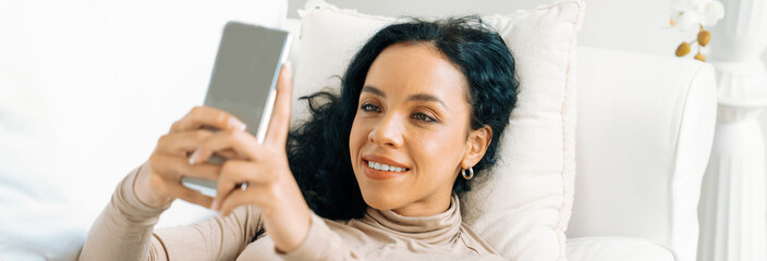 Relaxed young African American woman using crucial mobile phone on sofa couch in living room at home