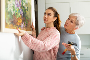 Young woman helping her elderly mother to hang painting on wall in room during arrangement of new...