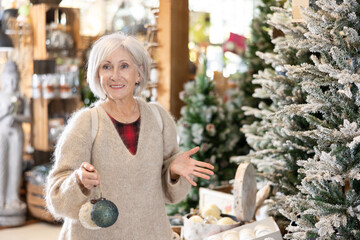 Elderly woman buyer examines toys for Christmas tree and imagines general style of house decorated for holiday
