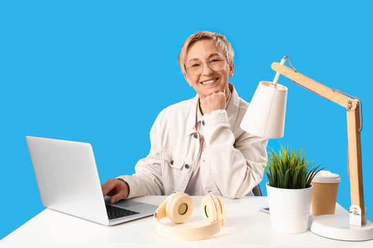 Mature female programmer with laptop at table on blue background