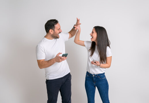 Ecstatic young couple giving high-five after reading good news over smart phones on white background