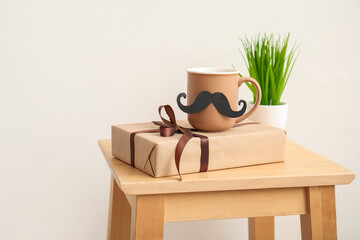 Cup of tea with mustache, gift and grass on table near light wall