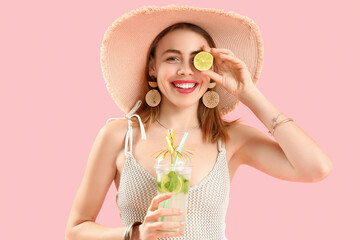 Beautiful young woman in wicker hat with glass of tasty mojito on pink background