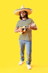 Handsome young Mexican man in sombrero playing guitar on yellow background
