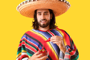 Young Mexican man in sombrero, poncho and with chili peppers on yellow background