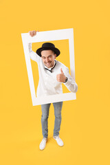 Happy young man with frame showing thumb-up on yellow background