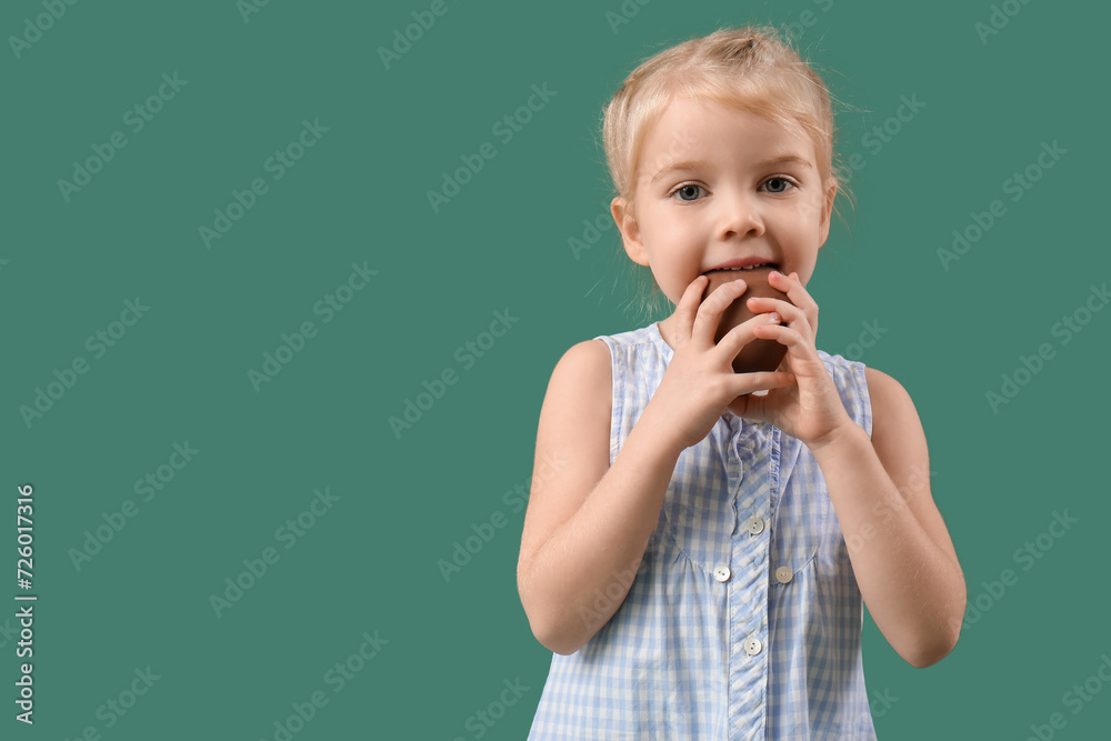 Wall mural Cute little girl eating chocolate egg on green background. Easter celebration