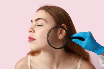 Dermatologist examining moles on face of young woman with magnifying glass on pink background,...