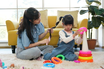 Asian mother braids her daughter's hair while sitting on the floor in the living room at home.