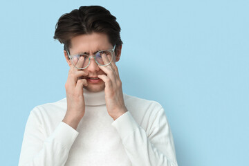Young man in glasses rubbing his eyes on blue background, closeup. Glaucoma awareness month