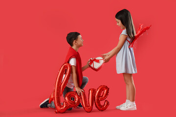 Cute little boy greeting girl with gift on red background. Valentine's Day celebration