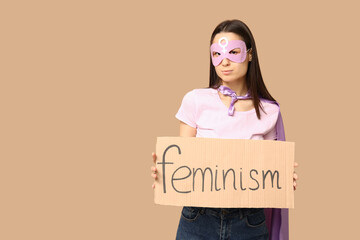 Woman in superhero costume holding cardboard with word FEMINISM on beige background