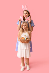 Young woman and her daughter in bunny ears holding basket of Easter cake with painted eggs on pink background